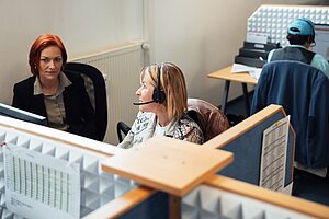 Zwei Telefonistinnen im Callcenter, eine Frau trägt ein Headset, die andere schaut aufmerksam zu. 