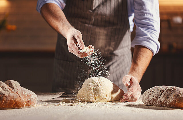 Ein Bäcker stäubt Mehl über einen frischen Brotteig um ihn zu formen und später zu backen.