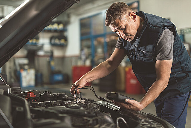 Das Bild zeigt einen Mechaniker, welcher unter einer offenen Motorhaube ein Auto repariert.