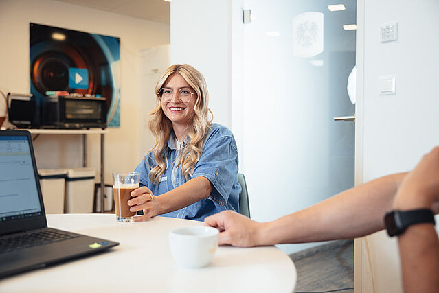 Mitarbeiterin lächelt und hält ein Glas Kaffee während einer Pause am Tisch, im Hintergrund ein Laptop und Büroeinrichtung.