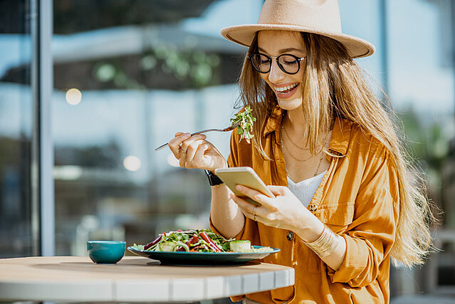 Junge Frau isst einen Salat und schaut sich Gastrotipps von FirmenABC auf ihrem Smartphone an.