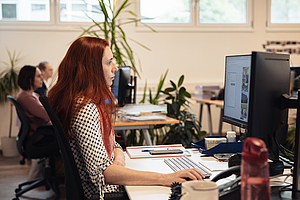 Frau mit roten Haaren arbeitet konzentriert an ihrem Computer, im Hintergrund weitere Arbeitsplätze und Pflanzen.